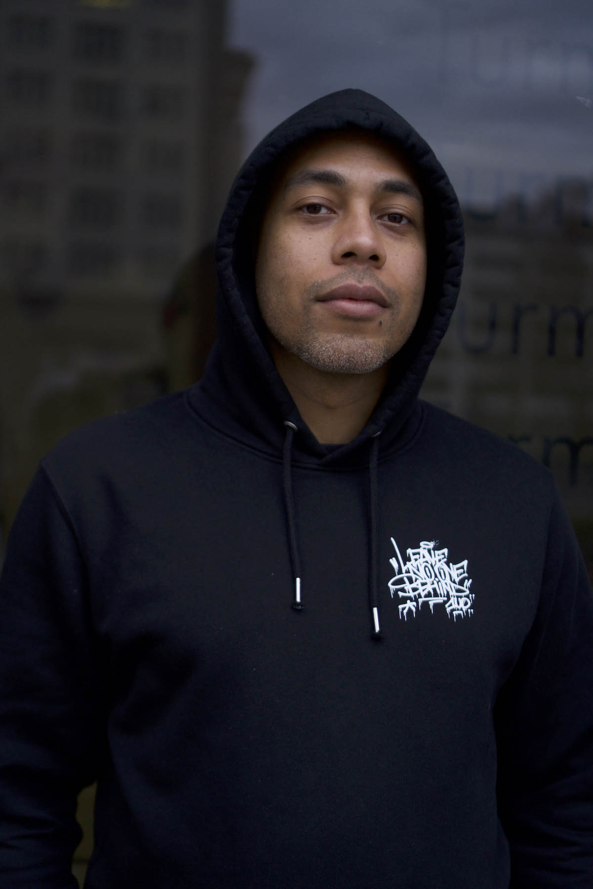 Black male person wearing a black hooded sweatshirt with the hood on his head looks at the camera.
