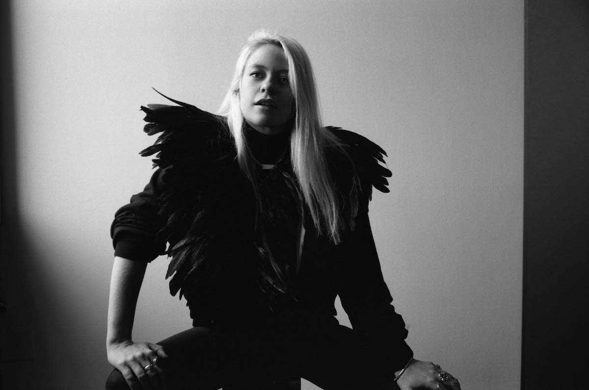 Black and white photo of blonde white woman with long hair. She can be seen up to her knees, sitting on a stool, resting her hands on her spread legs. She wears a black sweater trimmed with feathers, especially from her shoulders the feathers stick out to the side. She wears several rings on each hand and a septum piercing in her nose.