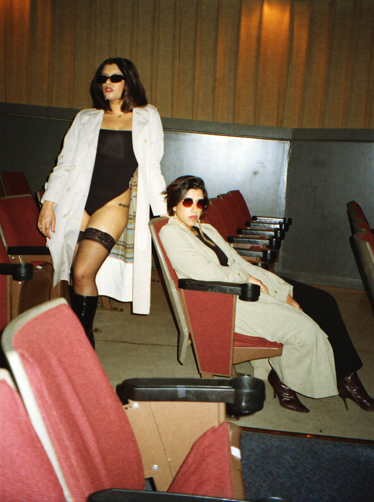 Two people with black hair in an old movie theatre, both wearing sunglasses. One of the two sits in a seat, this person wears black boots with spiked heels and a long sand-colored coat. The other is standing behind the seat, supporting herself with one hand on the backrest. This person is wearing a black bodysuit, black sheer overknee stockings with lace, black boots and a white coat.