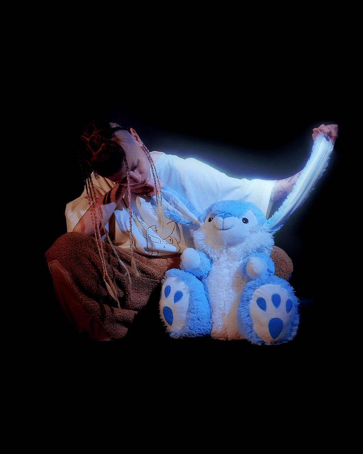 White female person sits on the floor in front of black background. She wears a white oversized shirt and brown plush pants. Her long hair is tied into several thin braids and shaved off at the sides. She is looking at a white and blue plush bunny sitting in front of her and lifting up its ears.