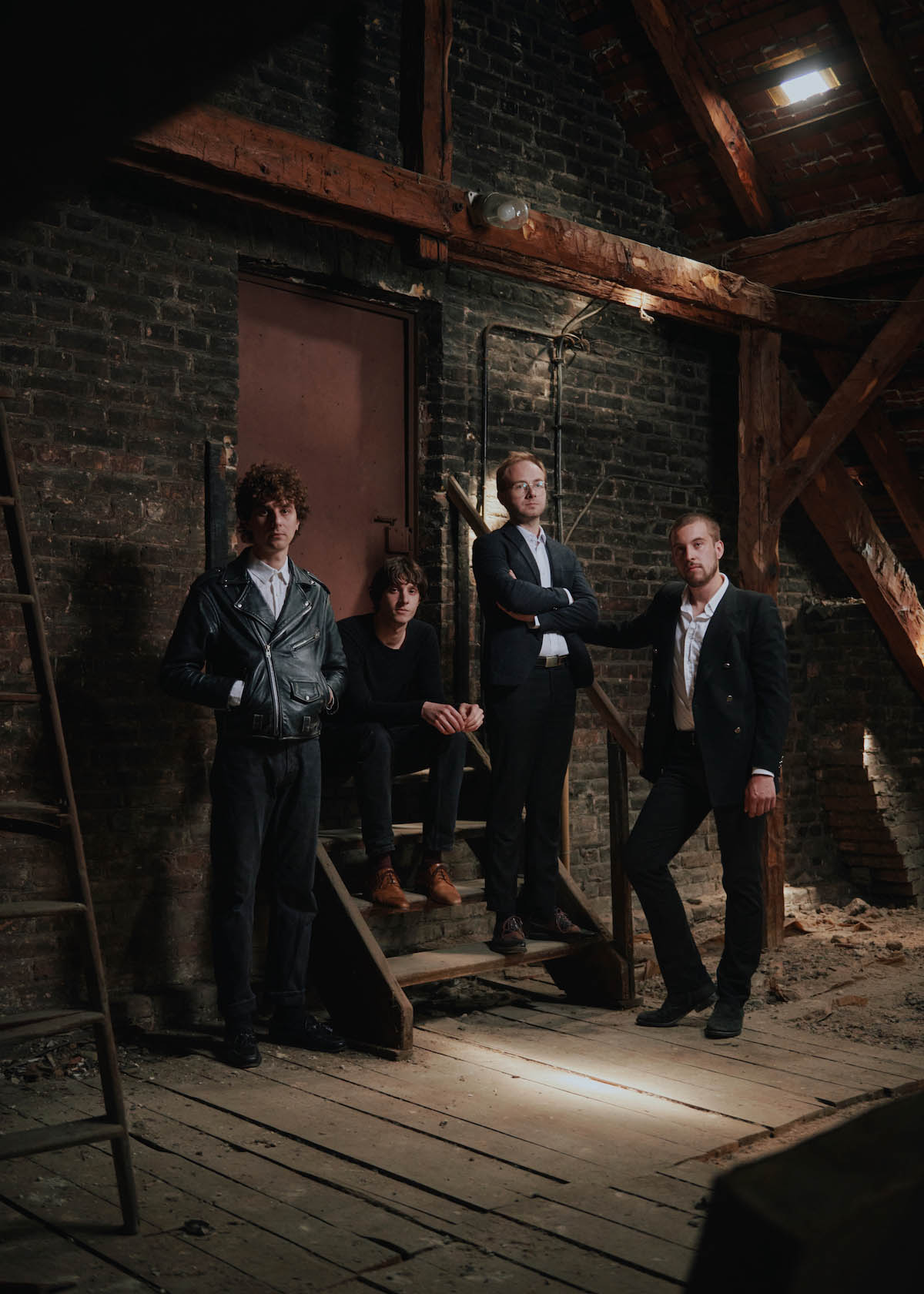 Four men look into the camera, three are standing up and around a four-step staircase in an attic-like environment, the fourth is sitting on this staircase. All of them are wearing black clothes.