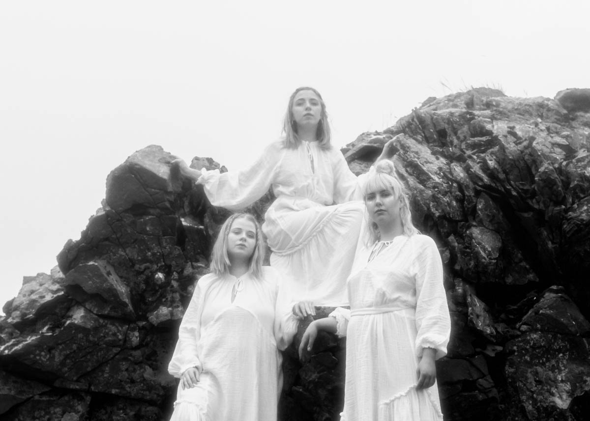Black and white photo of three blond women with shoulder-length hair in white robes. Two are standing in front of a rock, the third is squatting a little to the side between the two on the rock. They are all looking at the camera.