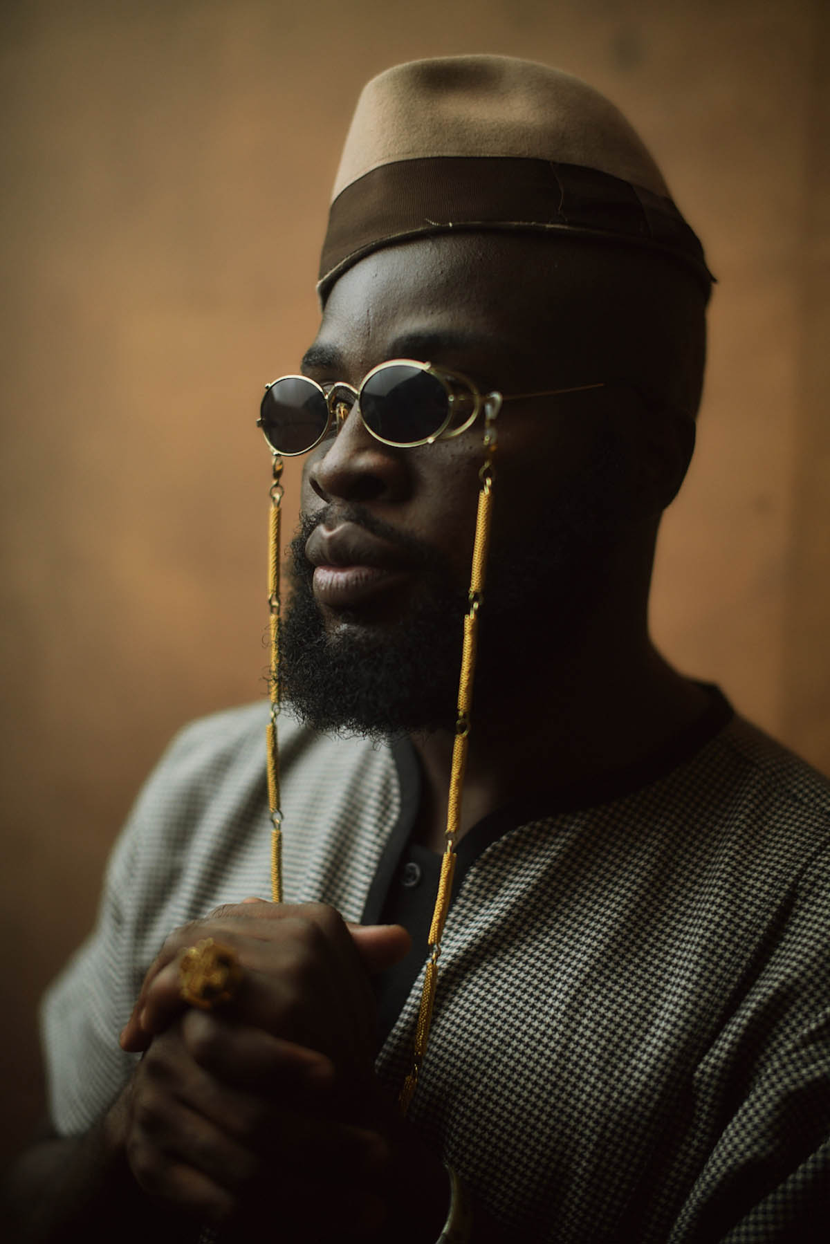 M.anifest is seen in a semi-close-up shot. He has his hands clasped together in front of his chest. He has a full black beard, wears sunglasses with gold frames and a yellow band hanging down next to his face. On his head he wears a hat without brim in light and dark brown.