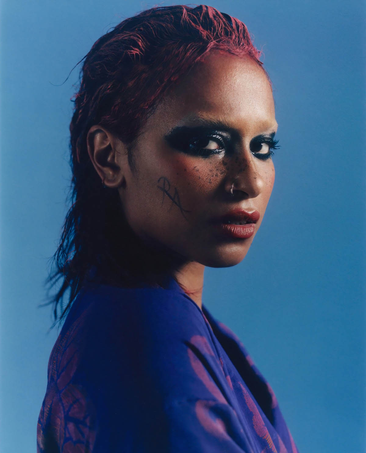 Female, Black person with darkly made-up eyes looks sideways into the camera. She wears a nose ring, a blue top and her red dyed hair is styled back. The letters "RA" are written on her cheek.