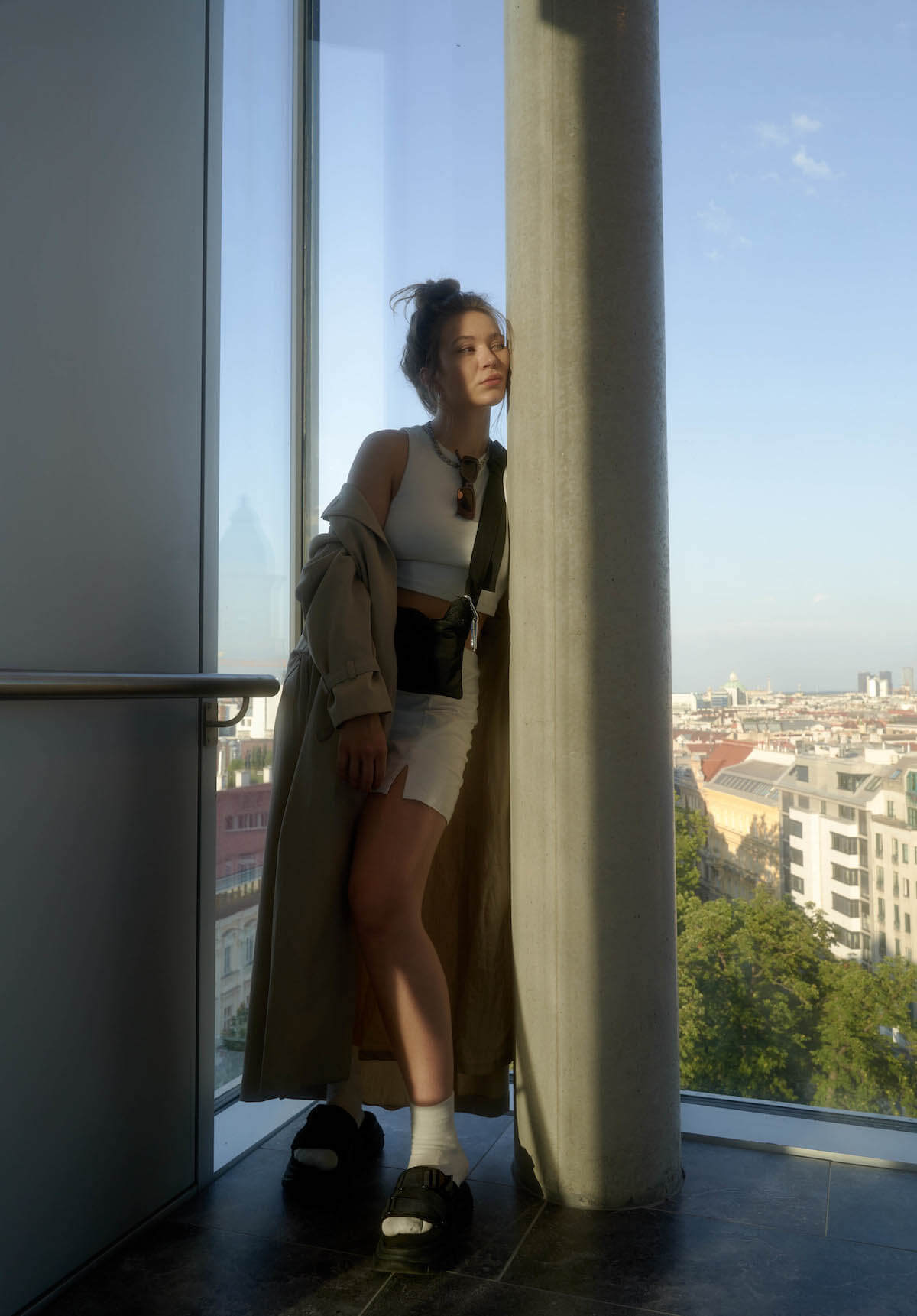 Young woman leaning against a column in front of a floor-to-ceiling window. Her long hair is tied up in a chignon, she wears a white crop top, a white short skirt, white socks in black sandals with high soles. She carries a shoulder bag from her right shoulder to her left hip and a long sand-colored coat that she lets fall off her shoulder. There are sunglasses hanging from the collar of her top. She looks to the right out of the picture.