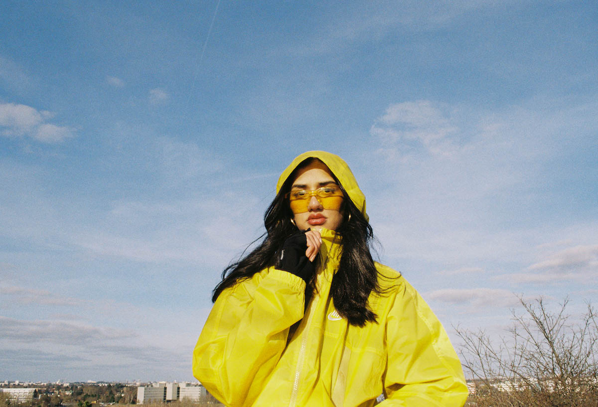 Young woman with long black hair and a yellow hooded rain jacket. She wears the hood on her head, her hair is falling forward over her shoulders. She’s shown down to her waist. In the background is mainly blue sky with white veil clouds. In the distance at the bottom of the picture you can make out high-rise buildings. Wa22ermann wears large yellow sunglasses that cover her face from her eyes to the tip of her nose. She looks into the camera and grips the zipper of her jacket with her right hand, as if she had just closed the jacket up to her chin.