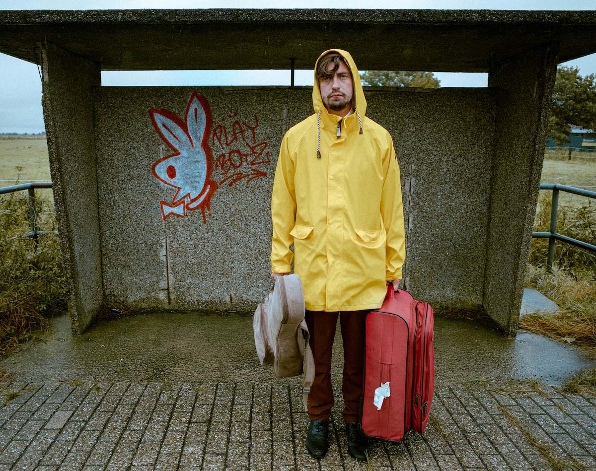 David Julian Kirchner, a white man with brown hair and a beard, stands in front of an abandoned bus stop in the countryside wearing a yellow mackintosh with the hood up. He looks into the camera. The ground is damp. Behind the concrete shelter, on which the graffito of a stylised Playboy bunny is spray-painted with the word "Playboyz", a field can be seen. In one hand Kirchner carries a red suitcase, in the other a beige guitar bag.