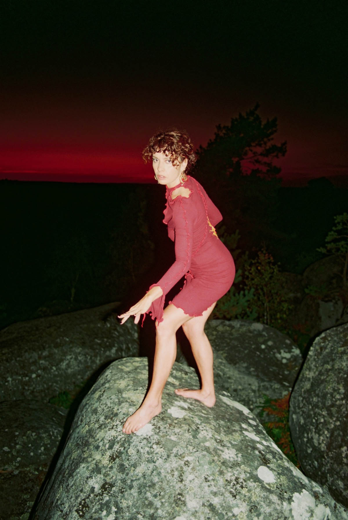 A white woman stands on a grey spotted rock with other bis rocks around her. It is Julie Bessard, alias HSRS. She is wearing a red, tight-fitting dress that reaches just above the knee, with long sleeves. Her legs and feet are bare. She has red, curly, pinned-up hair. She makes a slight lunge forward with her left foot, seemingly balancing herself with her left arm, which she delicately holds forward. The scene takes place at night, on the horizon the sky is dark red, all around is black, only the protagonist is illuminated by the flash of the camera.