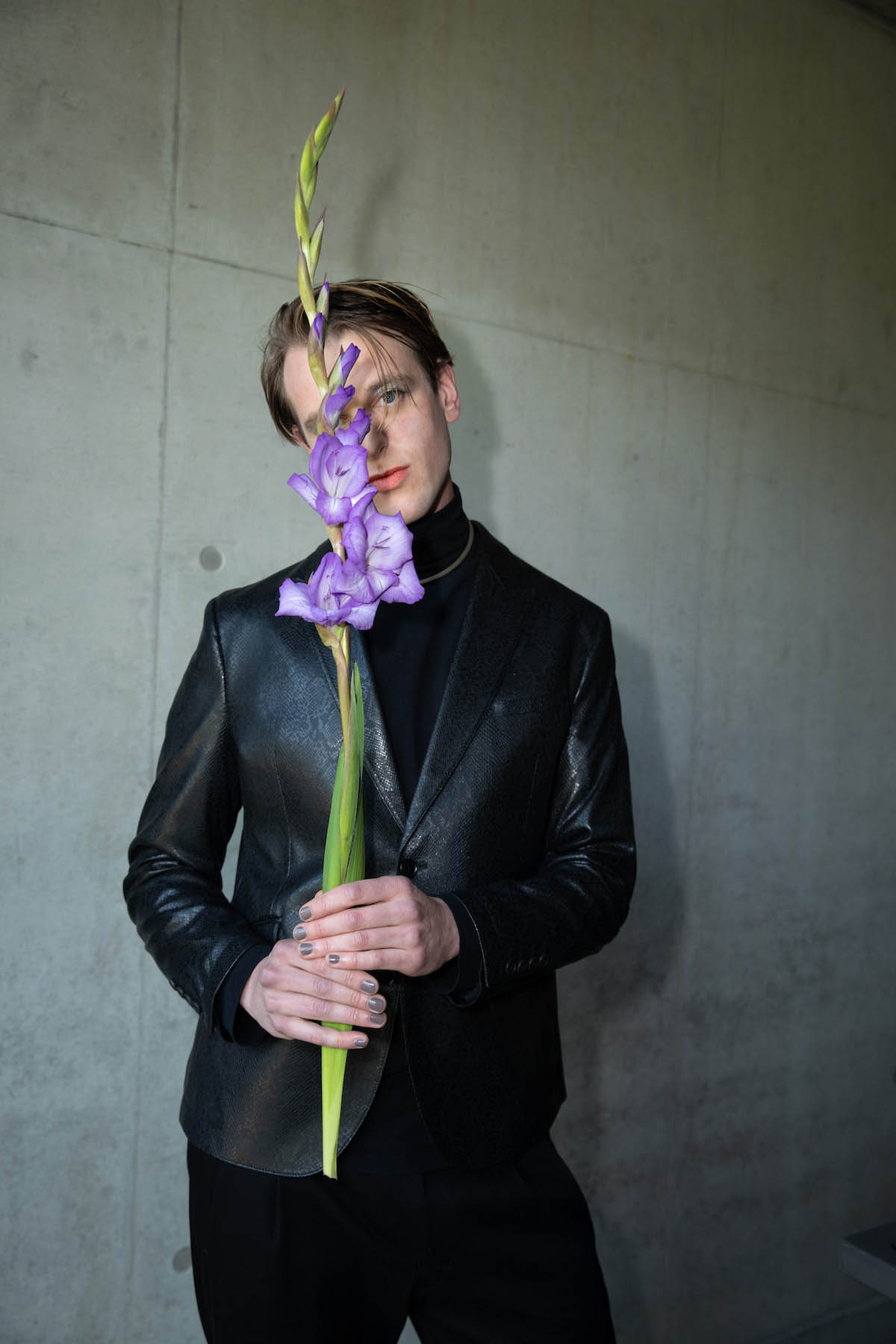A white, male person stands in front of a grey wall. He holds a long-stemmed flower with many purple blossoms in front of the face, covering half of it. Levin Goes Lightly looks into the camera, he is dressed completely in black, black turtleneck, black trousers, black leather jacket. Around the turtleneck he wears a beige-brown thin band. The colour returns on his nails. He holds his head at a slight angle. His hair is dark blond, a thin strand falls into his forehead.