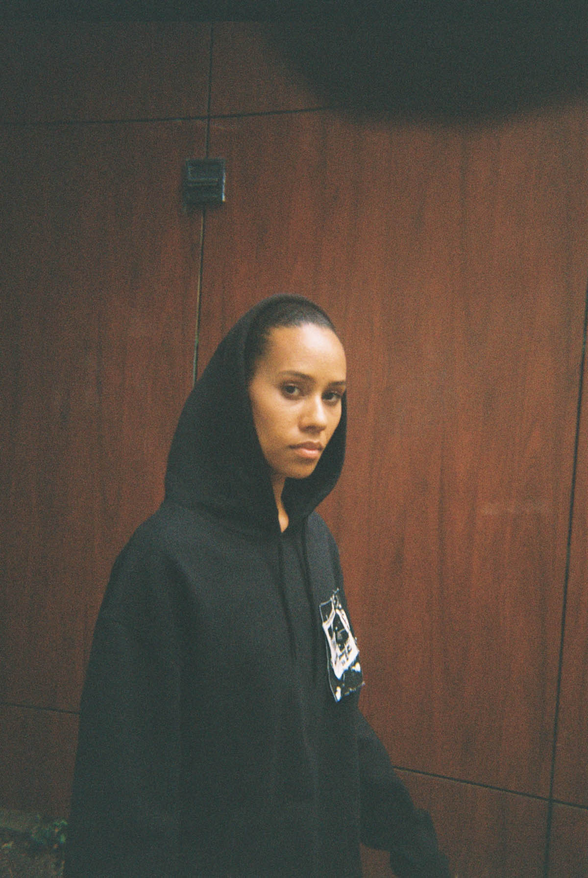 May The Muse can be seen up to her waist, she is standing at a slight angle to the camera and is wearing a black hoodie with a black and white large patch on the left chest. She wears the hood on her head and looks solemn. The part of her black hair that is still sticking out is combed back. The wall behind her is paneled with brown, large wooden panels. From above, a semicircular shadow protrudes into the picture.