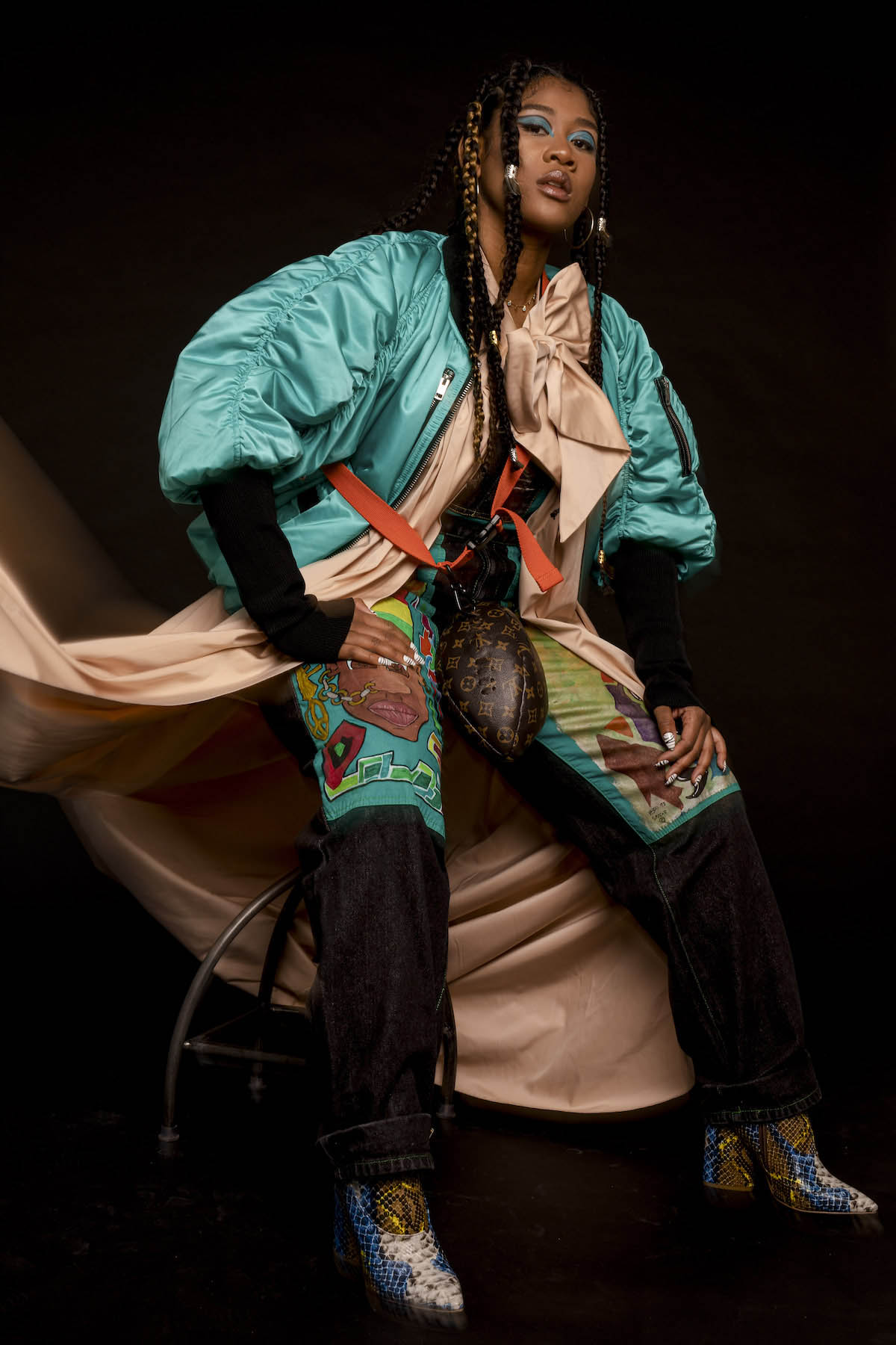 Rose May Alaba sits bent forward on a bar stool. Her legs are almost stretched out as the stool is high. She wears high-heeled, pointed shoes in a blue-beige-gold snakeskin look, black trousers with large turquoise-coloured patches covering her thighs. One of the patches shows the face of a black person with a chain from nose ring to ear. Between Rose May Alaba's legs hangs a brown bag with a red strap and typical Louis Vuitton pattern, the shape of which looks like an American football. Rose May Alaba rests her right hand on her right leg, the other rests loosely on her left. She looks firmly into the camera. Her eyelids are painted turquoise, the colour of her bomber jacket as well. Underneath, she wears an apricot-coloured long silk dress, open at the front, tied with a bow at the chest and falling almost to the floor behind the bar stool. On the left side of the picture, however, it is draped airily upwards. Rose May Alaba's long black hair is braided into several plaits.