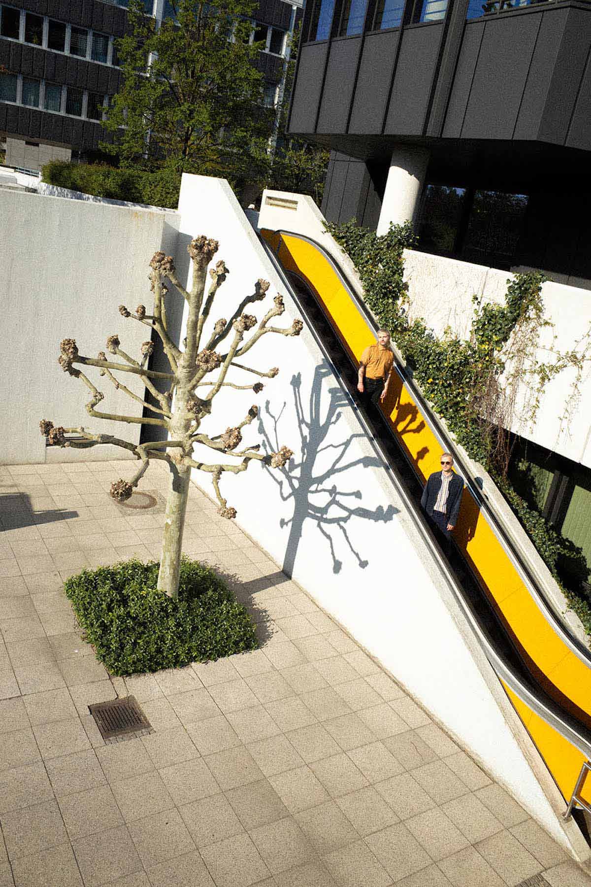 A yellow escalator leads down to a courtyard surrounded by office buildings with many windows. The courtyard is covered with square concrete slabs. In the courtyard, to the left of the escalator, a tree with thick branches and no leaves stands on a small square piece of green space. On the escalator, Schulverweis, two white men with short hair, can be seen on their way down, looking at the camera from a distance.