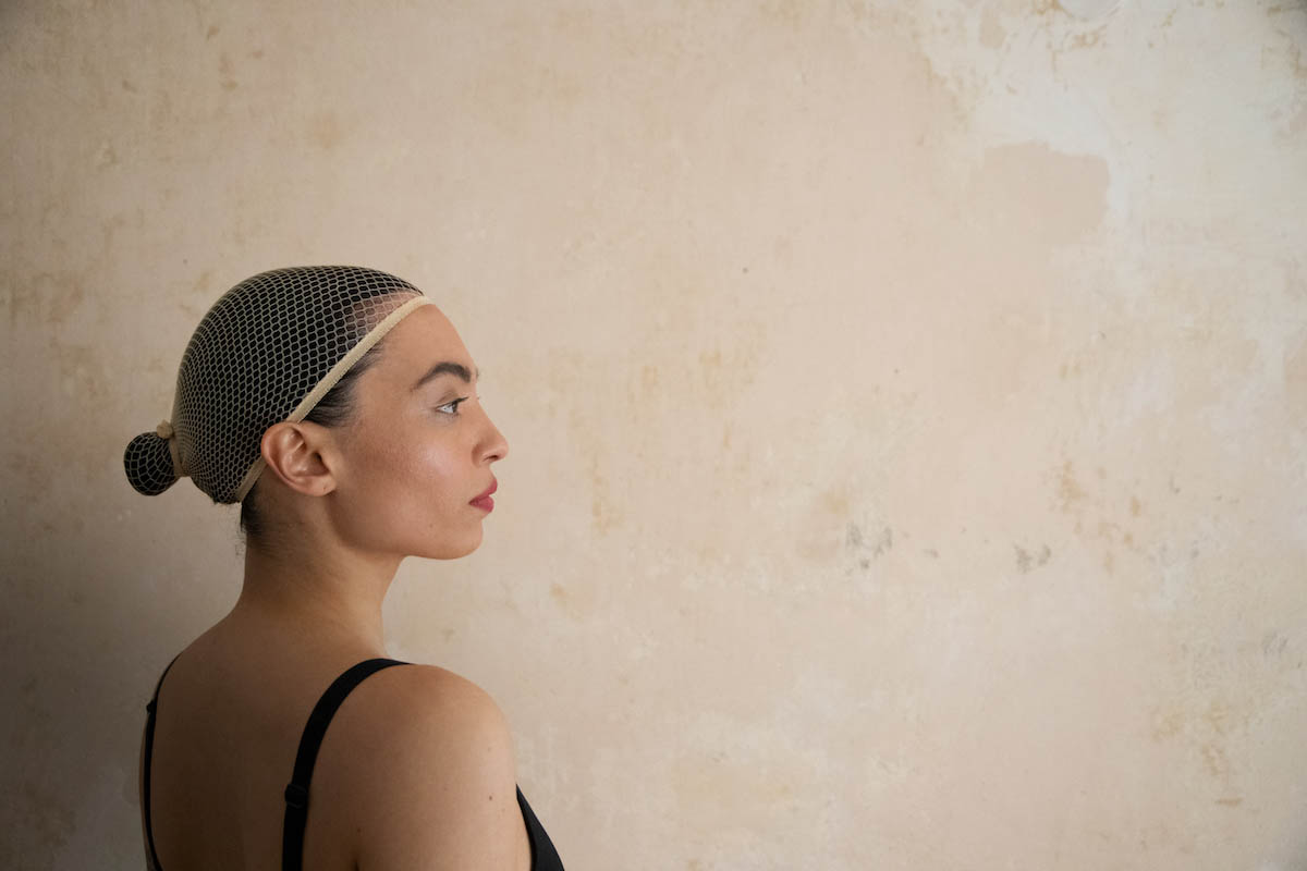 Vaovao has her dark hair strictly combed back and wears a chignon at the back of her head. Her hair is completely covered by a beige hairnet. She stands in front of an unplastered, light-colored wall and looks to the right out of the picture. Her upper body can be seen from diagonally behind until just below her shoulder. She is wearing a black bra, of which the straps can be seen. Her back and shoulder are bare.