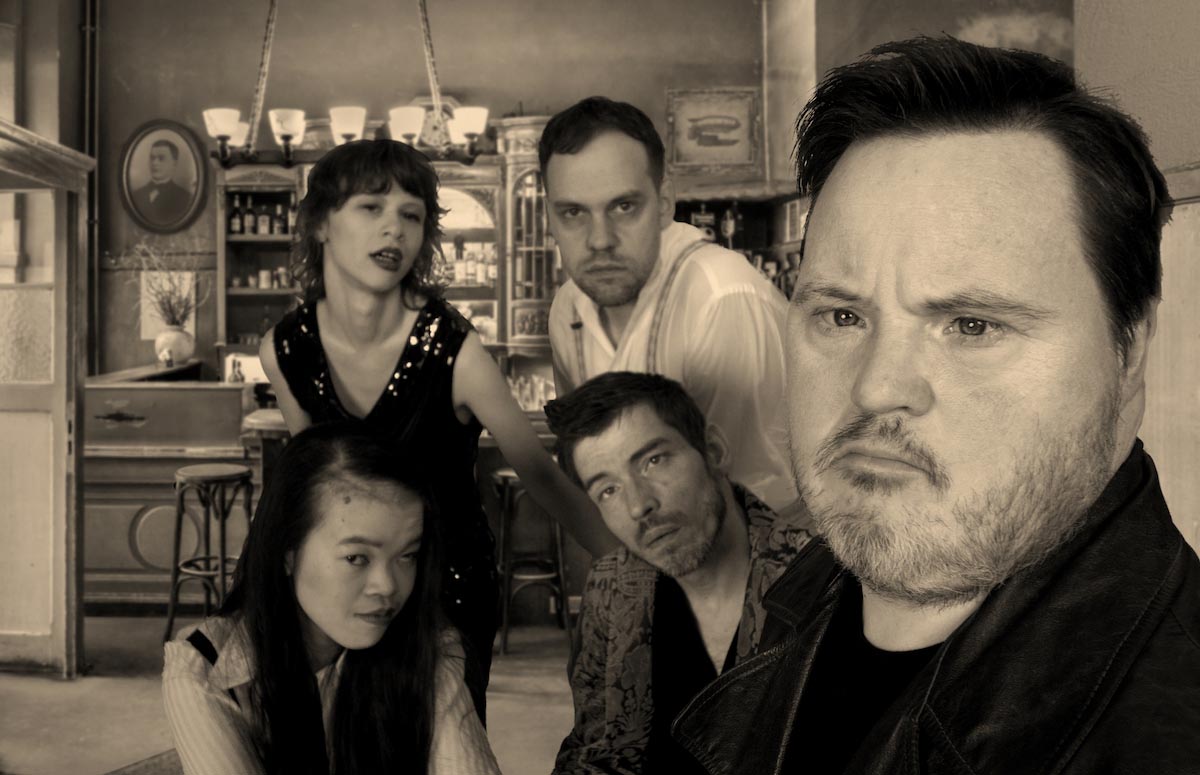 In the sepia-coloured picture of 21 downbeat, five people with and without visible disabilities - three men and two women - can be seen standing in front of an old-fashioned bar setting and posing for the camera. The person on the far right is closer to the lens, the others more in the background. All are looking at the camera.