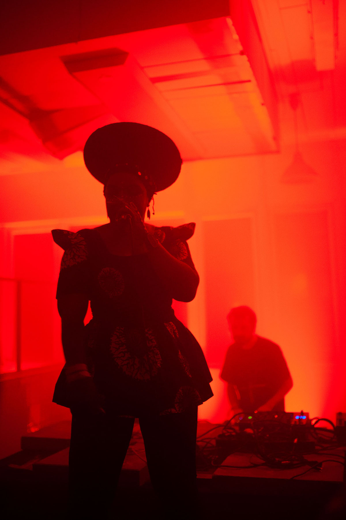 A performance situation with MC Yallah. She is standing in front of a mixing desk with many cables, a man can be glimpsed in the background. The stage behind her is illuminated by red light, which does not shine on MC Yallah. She stands in the dark and is not clearly visible. She wears what is called a Zulu hat, which fits tightly to her head and slopes upwards to finish in a large circle. She has a short dress with protruding shoulder appliqués and scattered large white flower-like patterns. With her left hand she holds the microphone in front of her mouth.